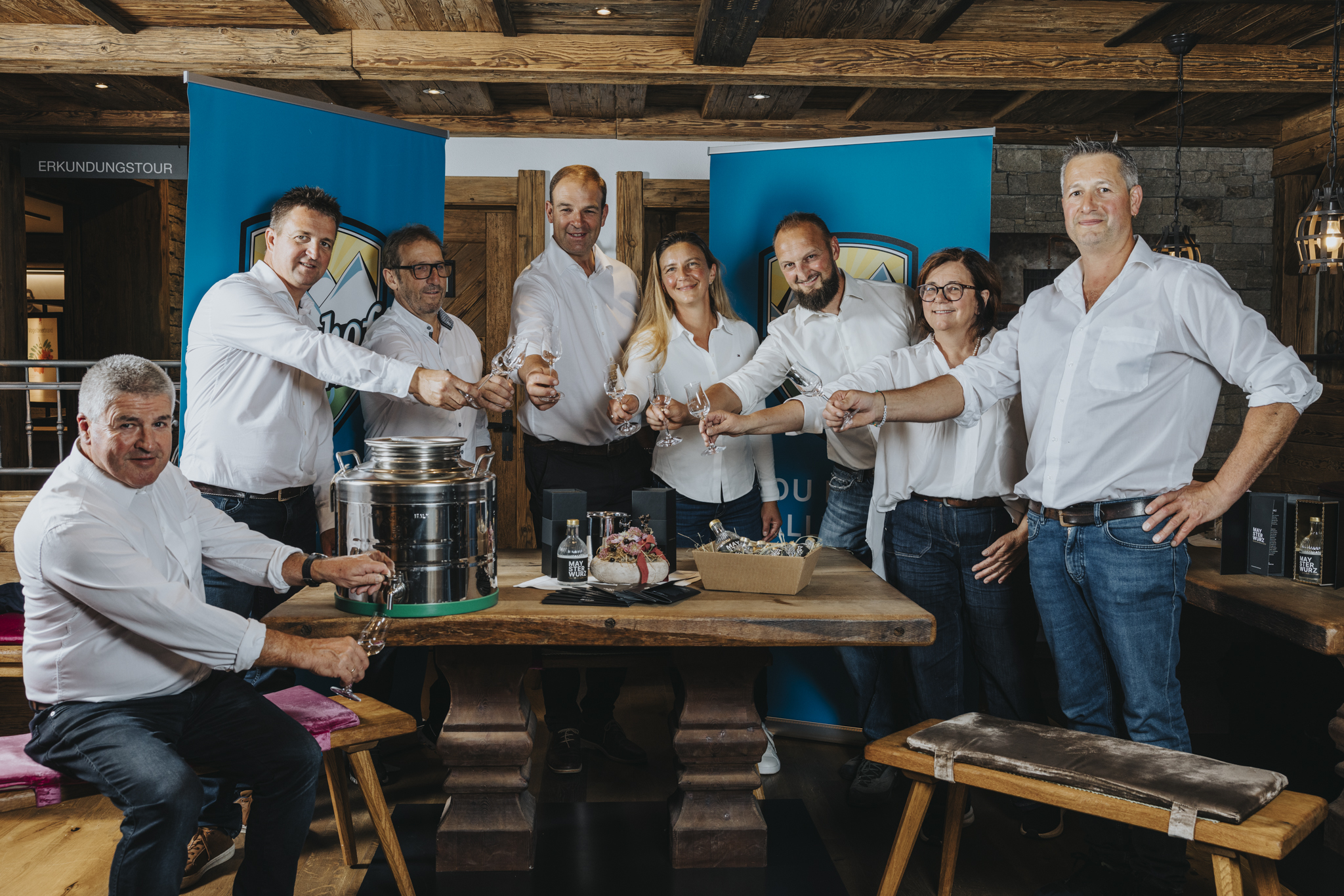 Acht Brennmeister des Maysterwurz stehen gemeinsam um einen Holztisch, heben ihre Gläser und stoßen in geselliger Runde an. Auf dem Tisch stehen Flaschen und Gläser zur Verkostung.