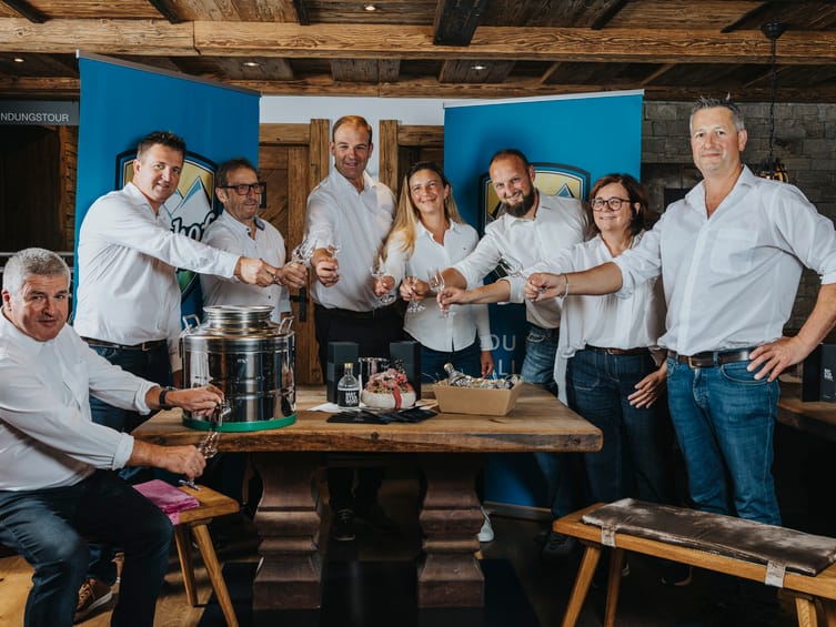 Acht Brennmeister des Maysterwurz stehen gemeinsam um einen Holztisch, heben ihre Gläser und stoßen in geselliger Runde an. Auf dem Tisch stehen Flaschen und Gläser zur Verkostung.