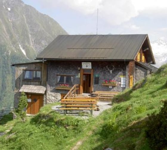 A mountain cabin with wooden stairs, flower boxes, and green grass in front of a mountain backdrop.