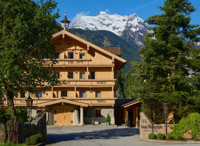 Gebäudeansicht des Hotel Gut Stiluppe mit 2 Bäumen links und rechts und im Hintergrund schneebedeckte Berge