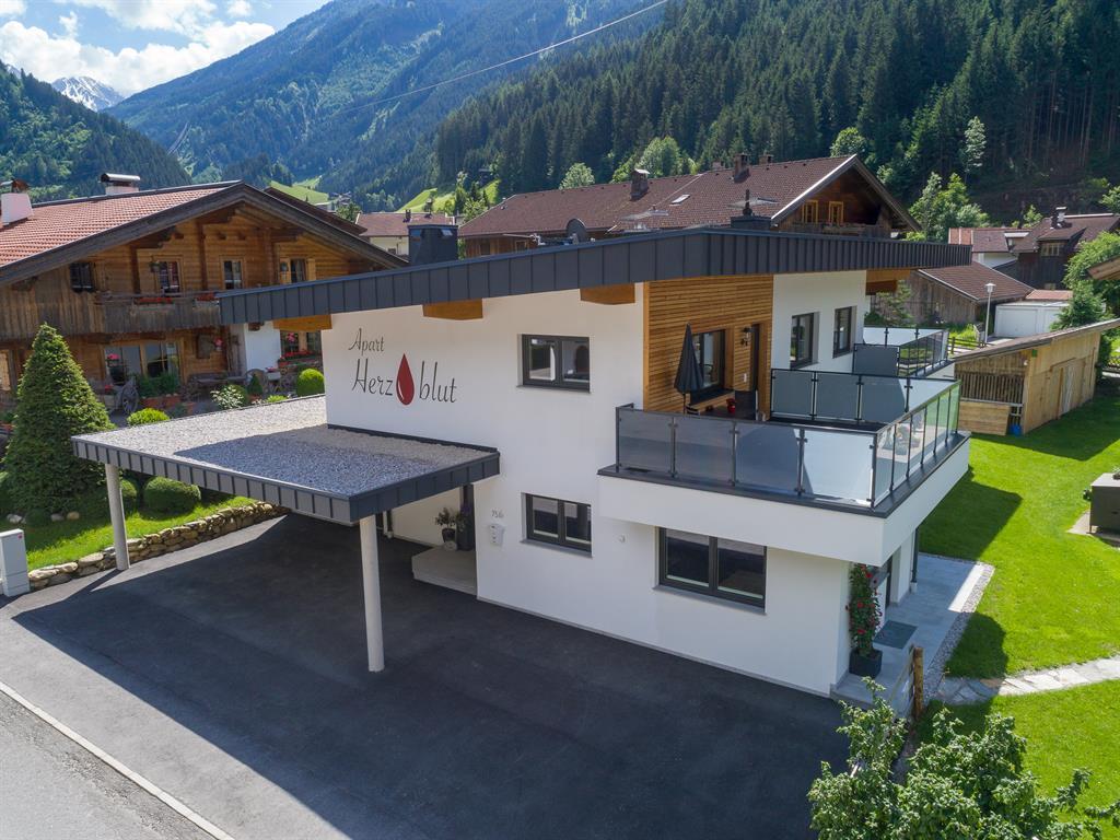 Modern house with balcony, in mountain scenery, with garden and neighboring houses in the background.