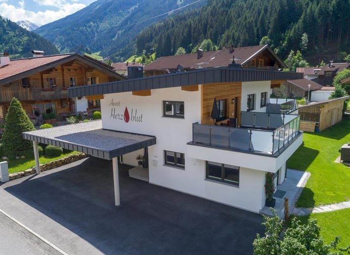Modern house with balcony, in mountain scenery, with garden and neighboring houses in the background.