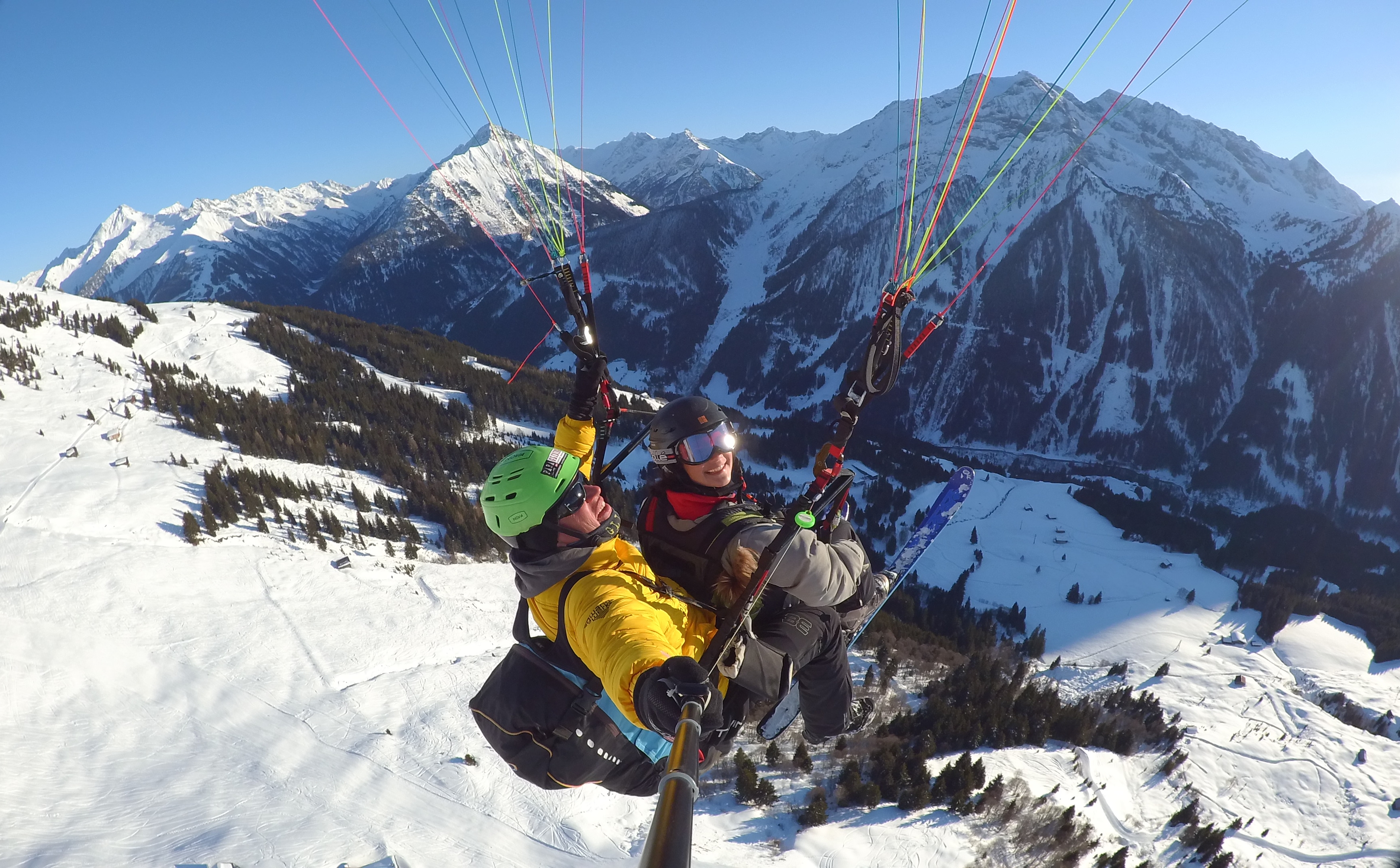 Zwei Skifahrer beim Paragliding in verschneiter Berglandschaft, mit Bergen im Hintergrund, sonnigem Himmel.