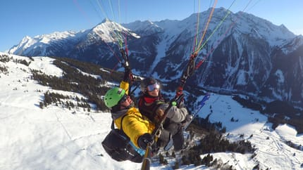 Zwei Skifahrer beim Paragliding in verschneiter Berglandschaft, mit Bergen im Hintergrund, sonnigem Himmel.