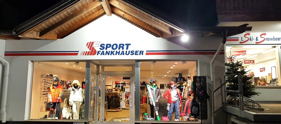 Eingangsbereich von Sport Fankhauser in Hippach bei Nacht, hell beleuchtetes Sportgeschäft mit Glasfront, Kleidungsausstellung und Treppe rechts – Skiverleih in der Ferienregion Mayrhofen-Hippach im Zillertal.