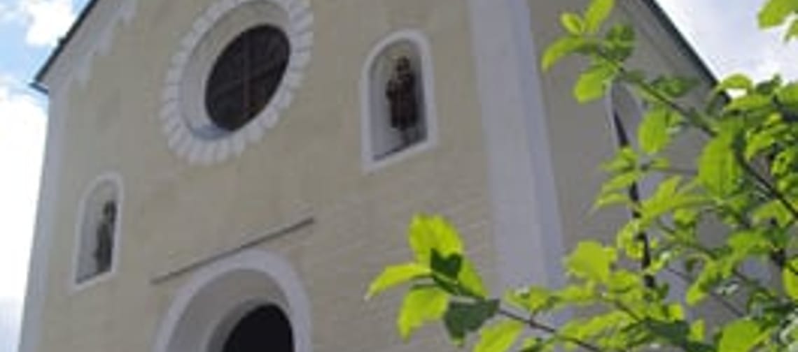 The Ginzling parish church is yellow with white stripes along the edges. The entrance and the cemetery are located in front of the church. To the right, you can see a green bush.