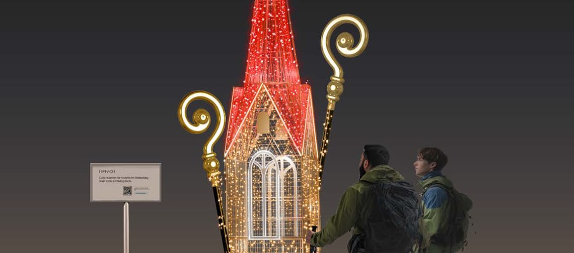 Beleuchtete Kirchturm-Installation bei Nacht in Hippach, Tirol: spitzer roter Turm mit goldenen Lichtornamenten auf rundem Sockel; zwei Personen mit Rucksäcken betrachten die Lichtskulptur vor dunklem Himmel.