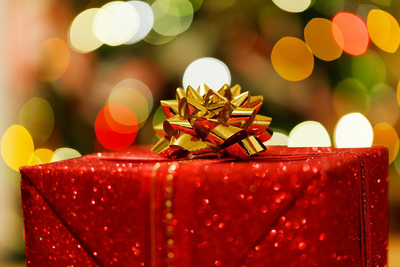 A glittering red gift wrapped with a golden bow in the foreground. Blurry, warm lights in the background create a festive Christmas atmosphere.