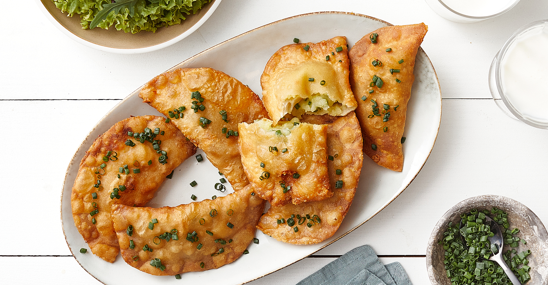 Top view of several golden-brown fried Zillertal Krapfen on an oval plate. One is broken open to show the potato and cheese filling. Sprinkled with fresh chives on a light wooden background.