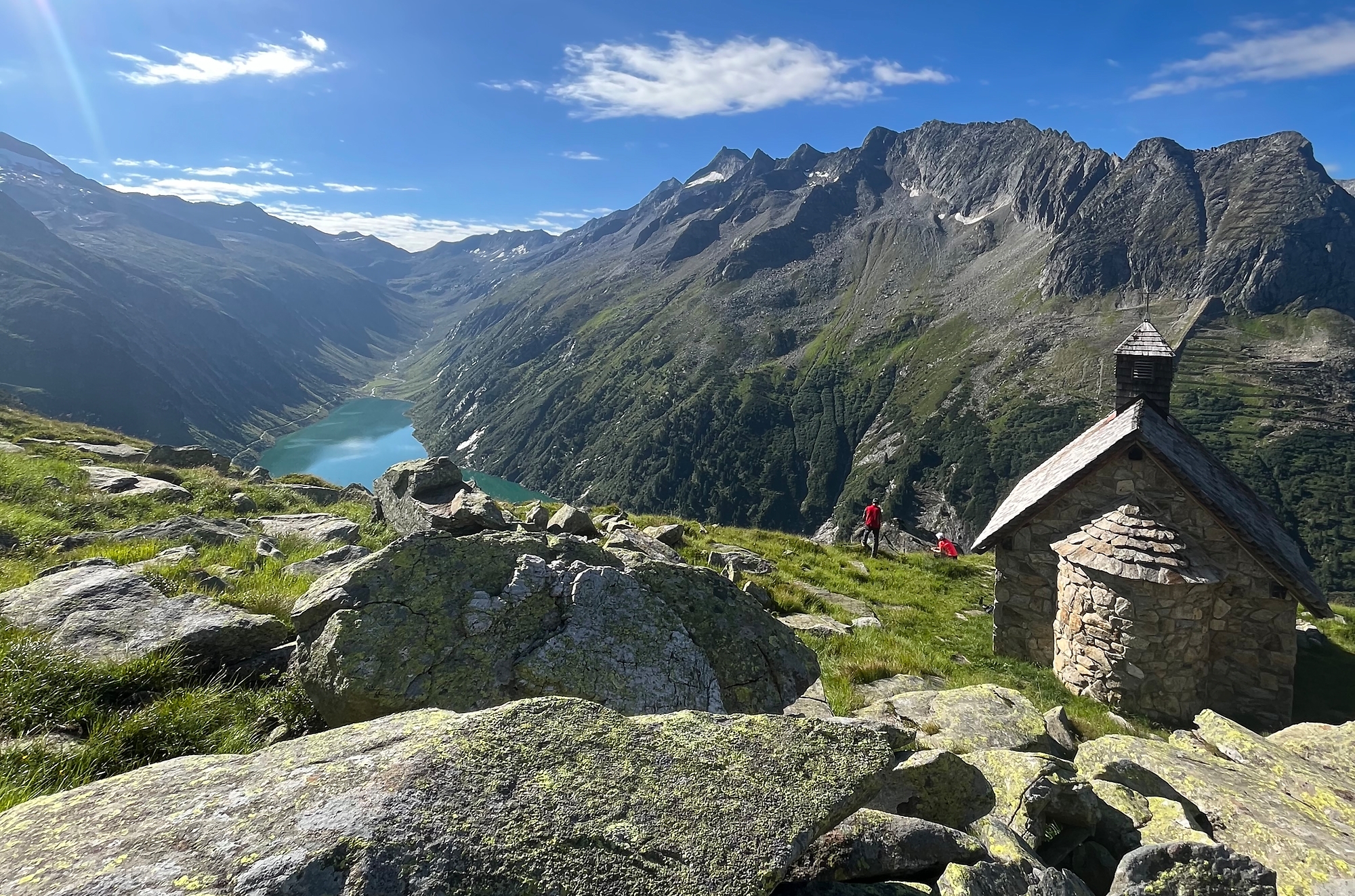 Beim Wandern im Zillergrund blickt man auf die kleine Kapelle zum Hl. Valentin, umgeben von Steinplatten und Almwiesen. Im Hintergrund liegt der türkisfarbene Speicher Zillergründl, eingerahmt von steilen Bergen unter blauem Himmel.