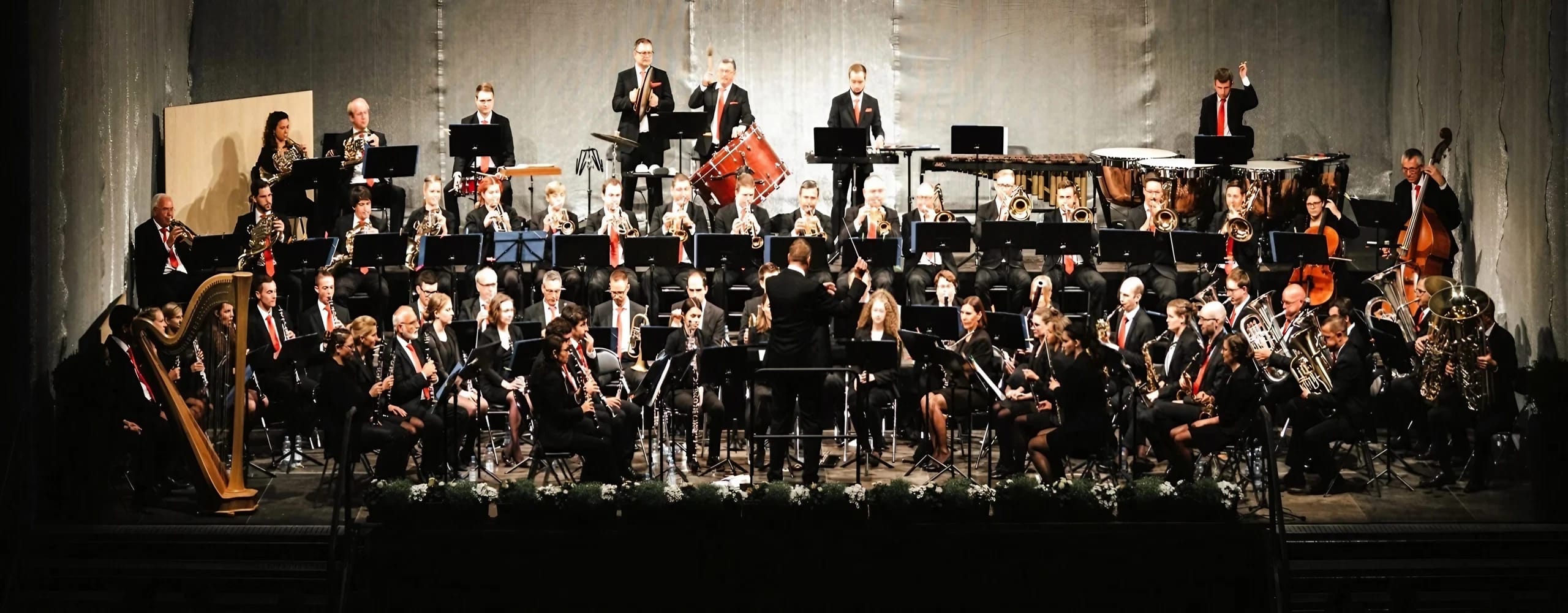 Großes Orchester auf einer Bühne. Viele Musiker in dunkler Kleidung sitzen mit Instrumenten, Dirigent steht vorne vor hellem Bühnenhintergrund.
