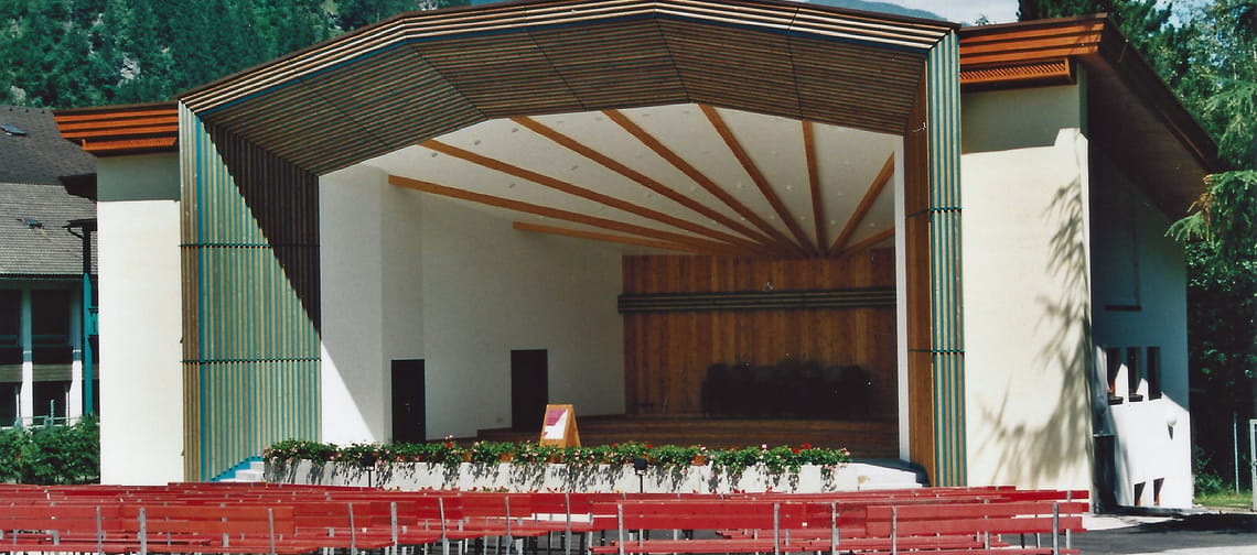 Historical color photograph of the music pavilion in Mayrhofen. The image shows the characteristic wooden stage construction and the red wooden benches set up for the audience.