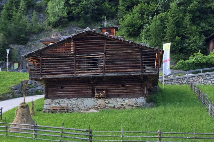 Mitterstall Brandberg Altes, zweistöckiges Holzhaus mit dunkler Fassade und steinernem Fundament auf grüner Wiese in Brandberg im Zillertal. Umgeben von Holzzäunen, mit bewaldeten Hügeln und Fahne im Hintergrund. Hier befindet sich die Ausstellung "Kulturlandschafts.Welten".