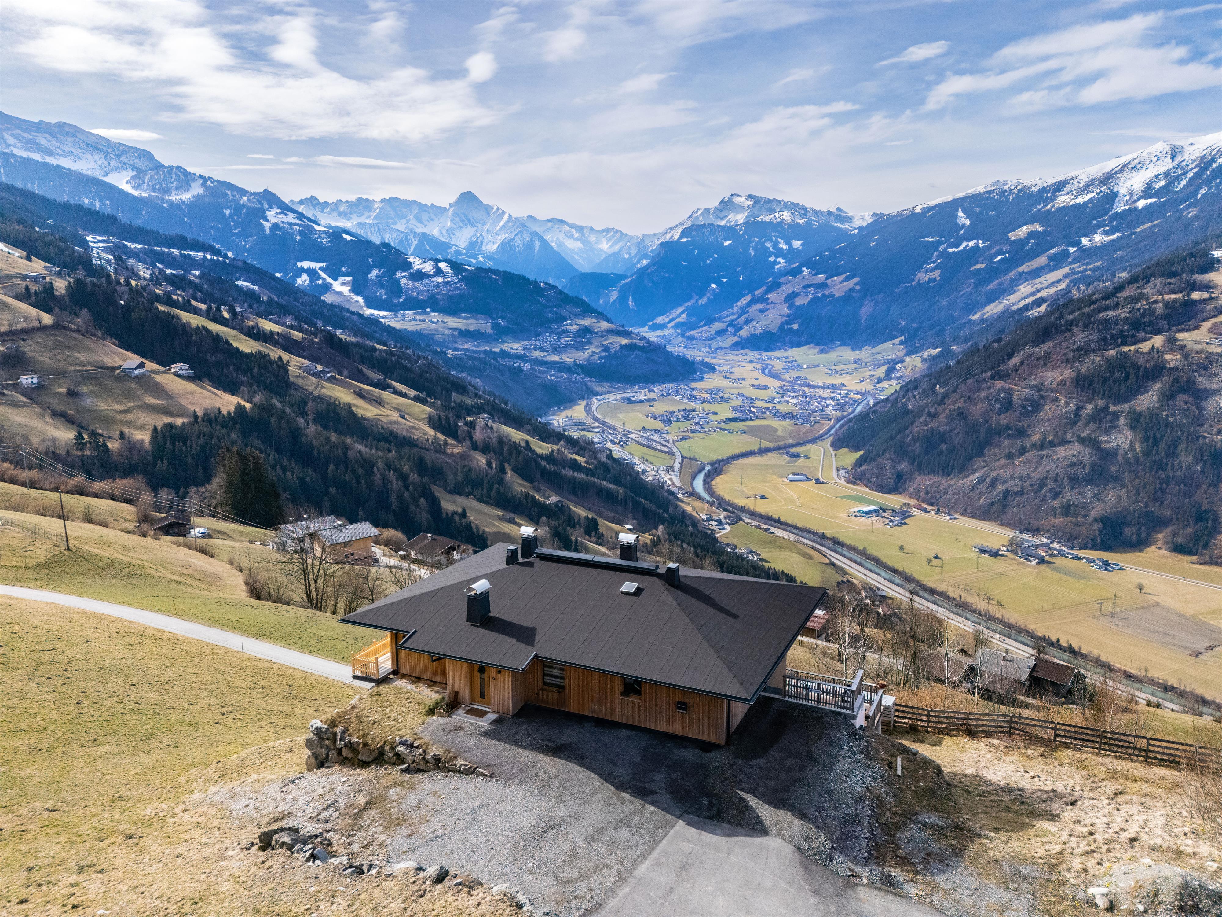 Houses in a mountainous landscape with fields, roads, and snow-capped peaks in the background.