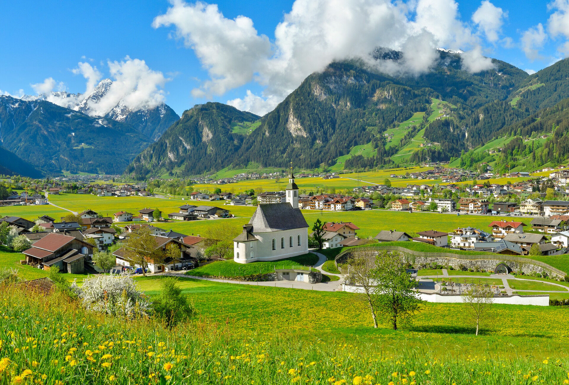 Ferienregion Mayrhofen-Hippach Blick auf das grüne Dorf Ramsau mit Wiesen, verstreuten Häusern und einer weißen Kirche im Zentrum. Im Hintergrund erheben sich bewaldete Berge unter blauem Himmel mit weißen Wolken.