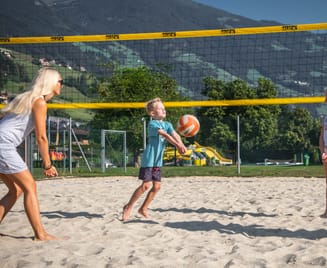 Familie beim Volleyballspielen auf dem Sandplatz der Sommerwelt Hippach, umgeben von Wiesen und Alpenlandschaft an einem sonnigen Tag.