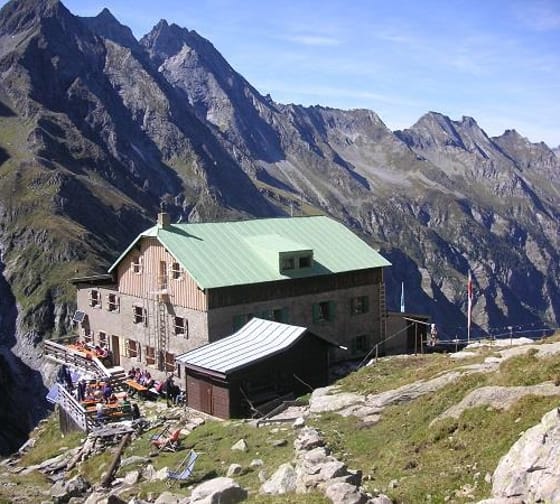 Mountain hut with green metal roof, surrounded by steep rocky peaks and a terrace with people.
