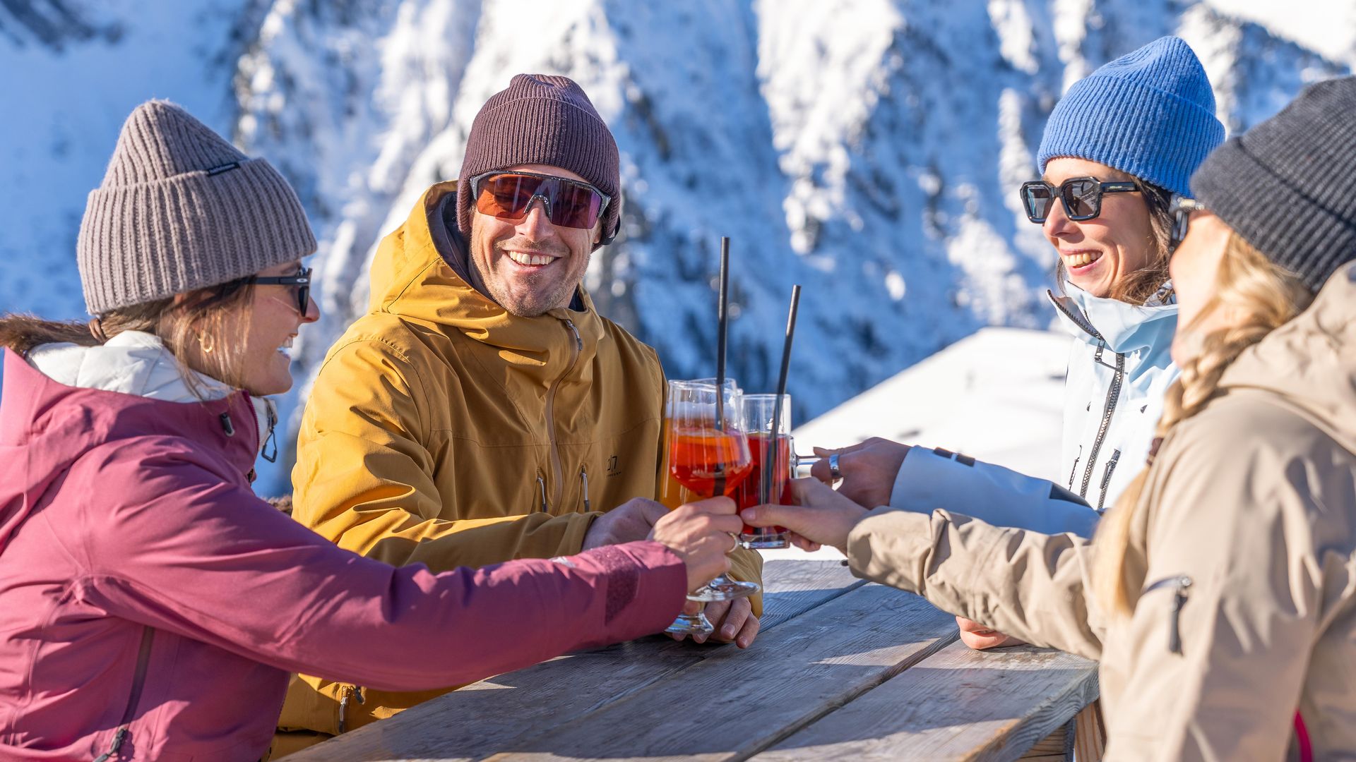 Gruppe von Menschen in Winterkleidung sitzt an einem Holztisch in einer Schneebar in Mayrhofen; sie unterhalten sich und trinken in der Sonne, im Hintergrund Schnee, Berge und blauer Himmel.