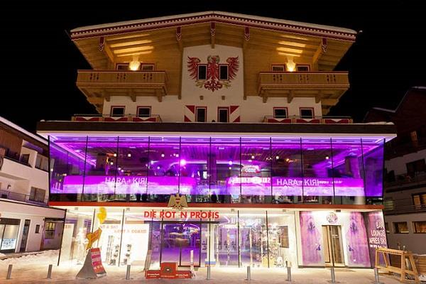 The image shows the Harakiri Bar from the outside at night. Pink LED lights shine from the second floor, giving the building a modern and striking appearance.