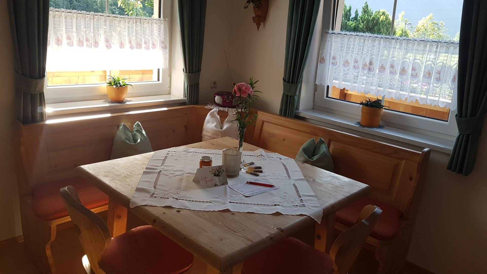 Ein Holztisch mit weißen Tischdecke, Blumen, Kerze und Medikamenten, umgeben von Fenstern mit Vorhängen, in einem hellen Raum.