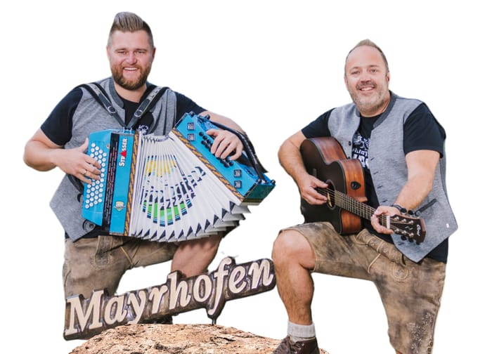 2 Männer stehen mit einem Fuß auf einem kleinen Felsen. Beide sind in Lederhosen und trachtigem Gilet gekleidet. Sie spielen mit Ziehharmonika und Gitarre.