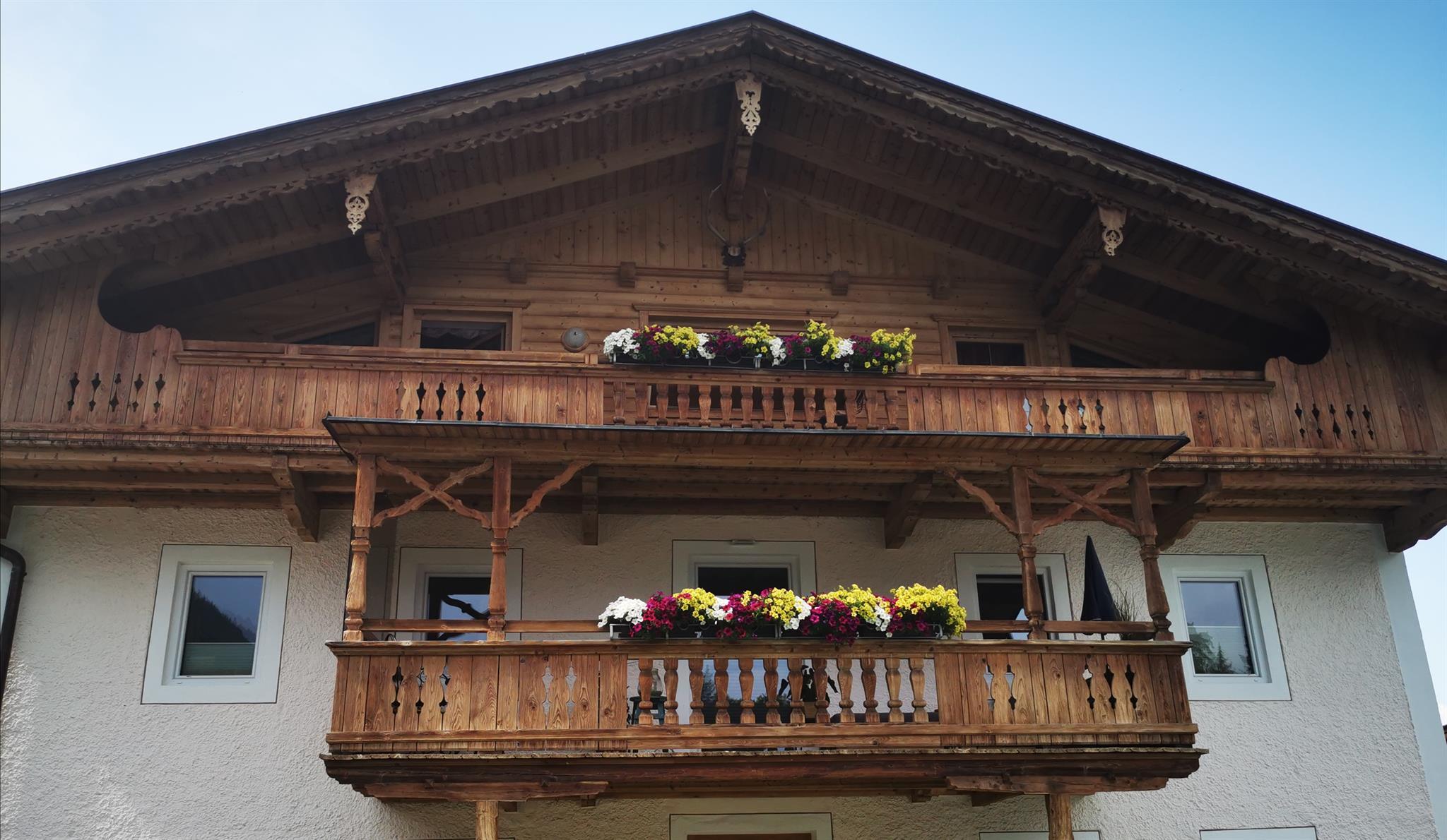 Mehrstöckiges Holzhaus mit Balkon, Blumen und Fenster, unter blauem Himmel.