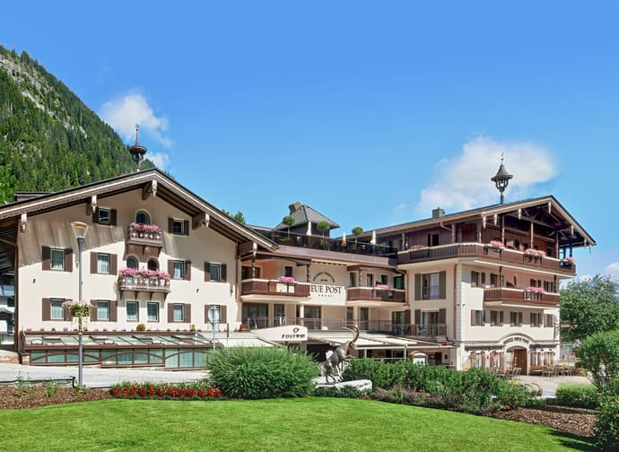 The image shows Hotel Neue Post from the outside, with a green meadow stretching out in front. In the background, a bright blue sky, scattered clouds, and majestic mountains create an idyllic setting