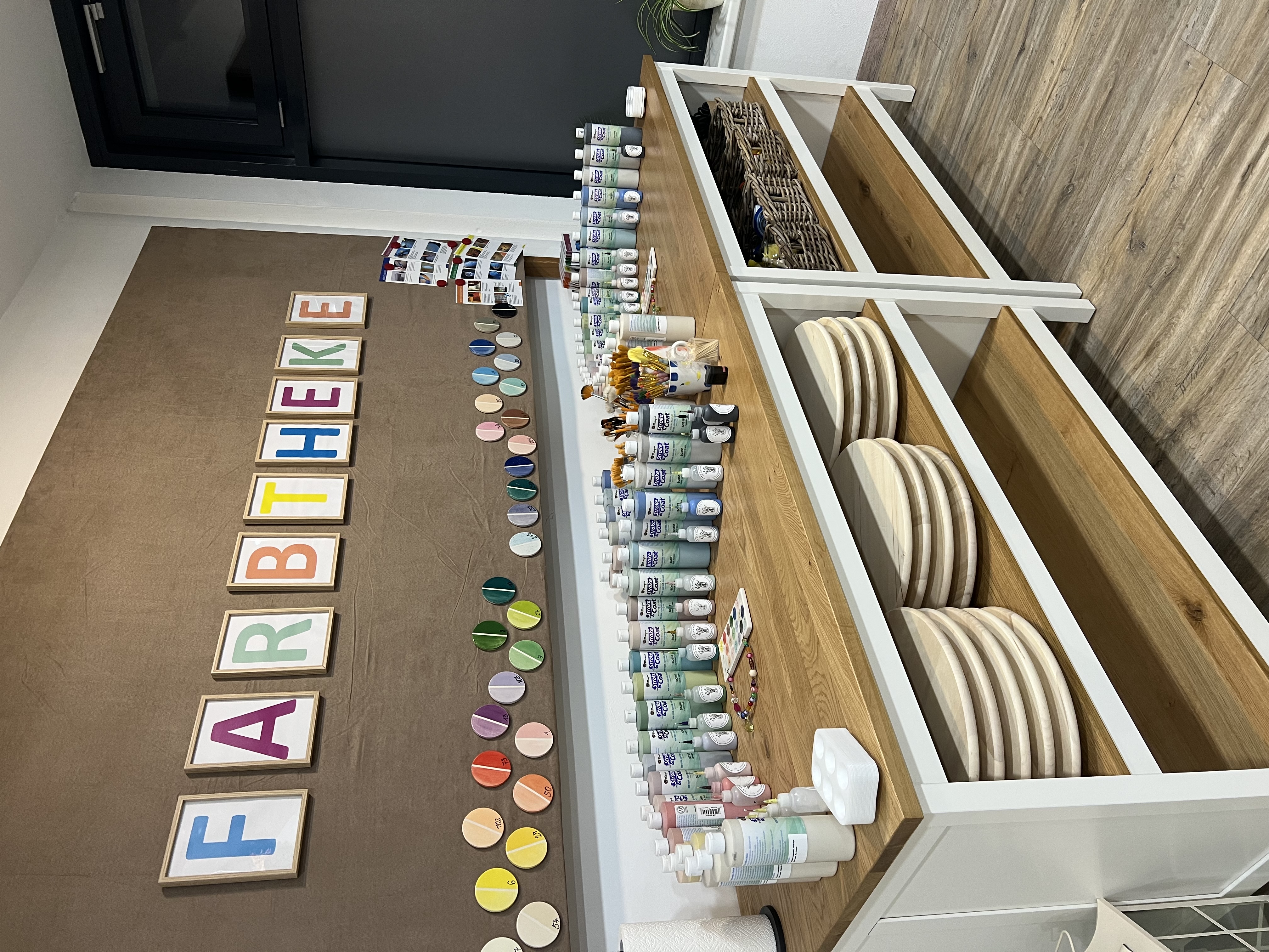 Ein Tisch auf dem verschieden Farbtuben stehen und ein Schriftzug mit "FARBTHEKE" an der Wand