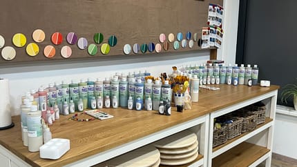 Ein Tisch auf dem verschieden Farbtuben stehen und ein Schriftzug mit "FARBTHEKE" an der Wand