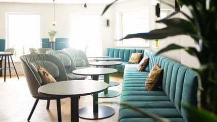 Das Bild zeigt eine gemütliche Sitzecke der Hof Bar – Wine & Cocktailbar mit einer türkisfarbenen Bank, passenden Sesseln und Stühlen. Im Vordergrund steht eine Pflanze, im Hintergrund sorgen große Fenster für eine helle und einladende Atmosphäre