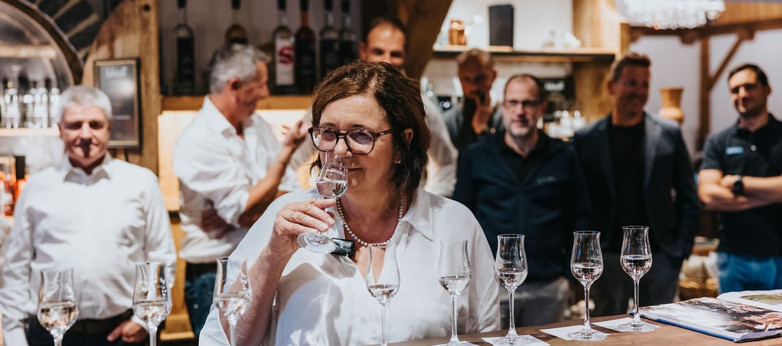 Das Bild zeigt die Ginin, die an einem Glas klaren Schnapses riecht. Vor ihr stehen mehrere gefüllte Gläser auf einer Trese, jeweils auf kleinen Untersetzern. Im Hintergrund stehen mehrere Männer, die dem Geschehen aufmerksam folgen.
