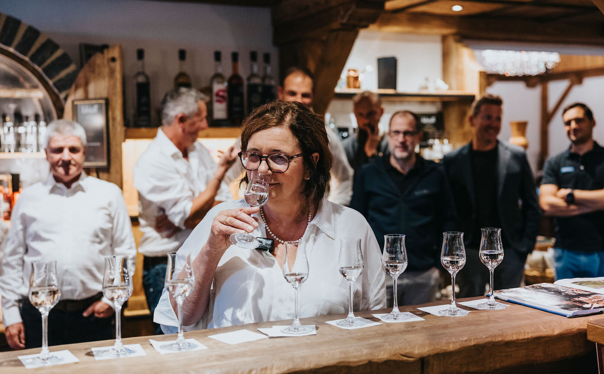 Das Bild zeigt die Ginin, die an einem Glas klaren Schnapses riecht. Vor ihr stehen mehrere gefüllte Gläser auf einer Trese, jeweils auf kleinen Untersetzern. Im Hintergrund stehen mehrere Männer, die dem Geschehen aufmerksam folgen.