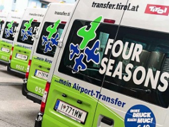 Station shuttle: Green Four Seasons shuttle buses at the airport with transfer.tirol.at lettering. The vehicles offer airport transfer services. License plates and company logos are clearly visible on the modern coaches.