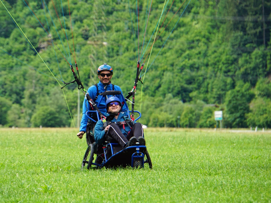 Rollstuhlgestützes Gleitschirmfliegen - Landung in Mayrhofen Ein Tandem-Gleitschirmflug mit Rollstuhlunterstützung landet auf einer grünen Wiese. Der Passagier sitzt lächelnd im speziellen Rollstuhl, während der Pilot hinter ihm steht.