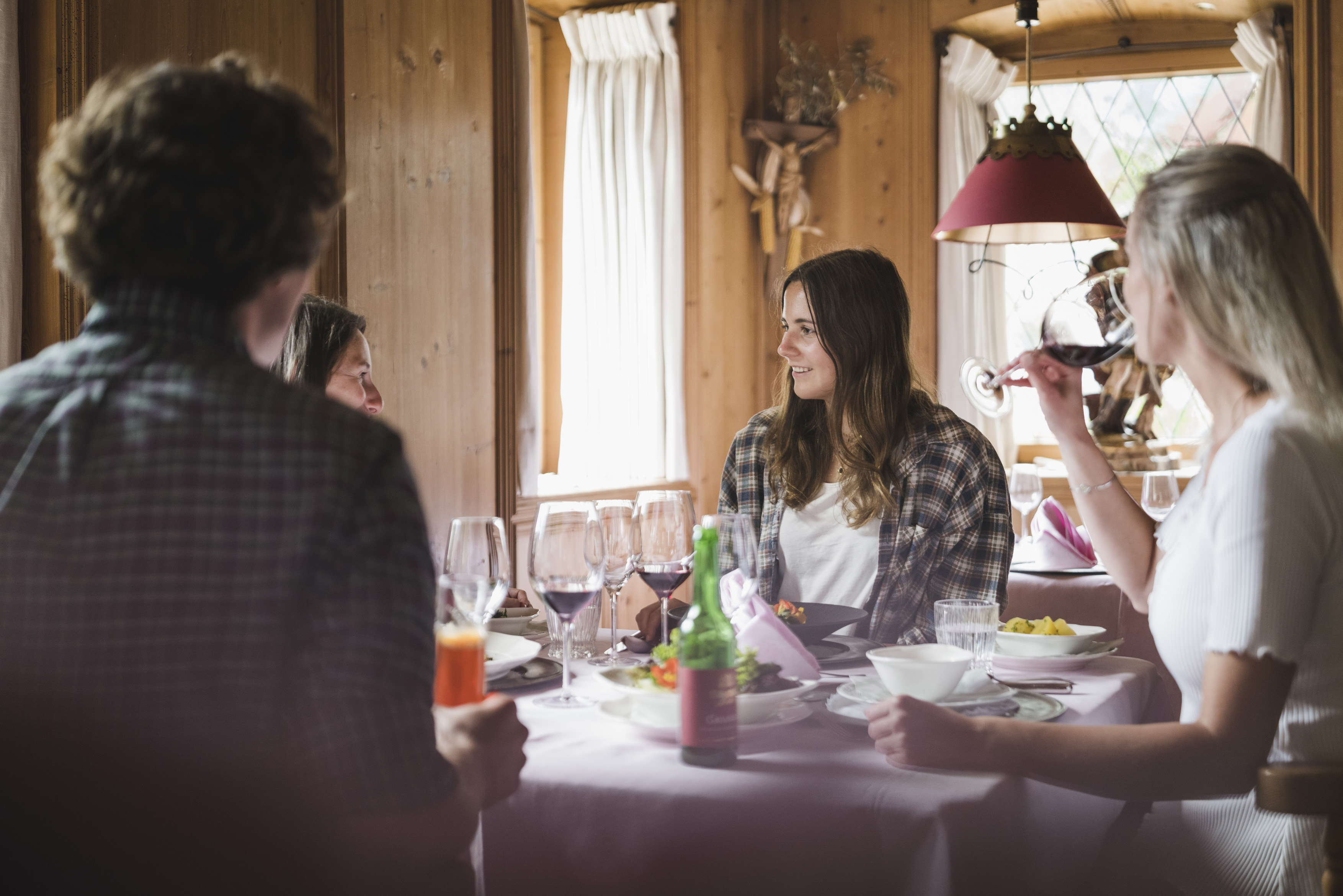 Eine Gruppe von Menschen sitzt beim Essen an einem festlich gedeckten Tisch in einer hellen, holzgetäfelten Zirbenstube im Hotel Neuhaus. Eine Frau trinkt ein Glas Rotwein, während im Hintergrund die traditionelle Einrichtung für Gemütlichkeit sorgt.