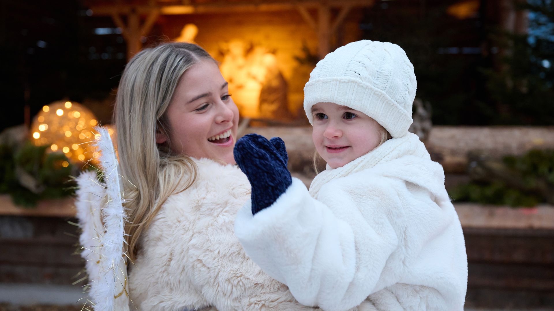 A woman in a white fur coat carries a child dressed in white winter clothes and a hat. Both are smiling, with warm Christmas decorations glowing in the background.