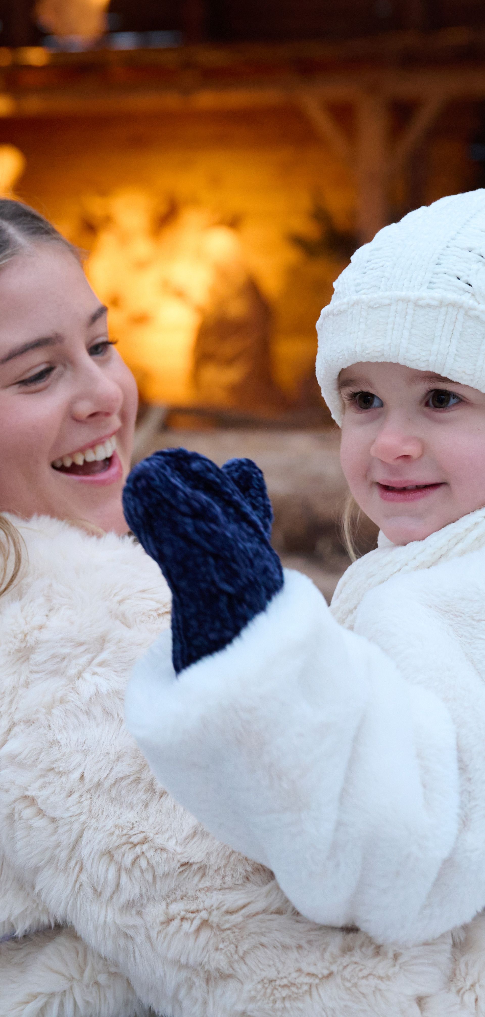 A woman in a white fur coat carries a child dressed in white winter clothes and a hat. Both are smiling, with warm Christmas decorations glowing in the background.