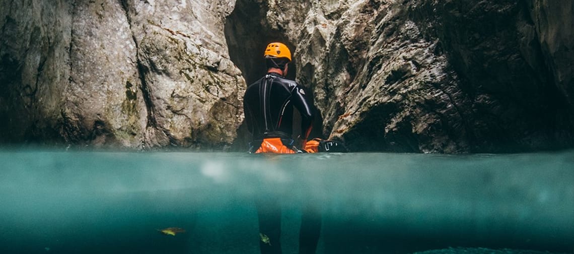 Das Bild zeigt einen Mann, der mit den Beinen im glasklaren Wasser steht. Vor ihm sind Felsen zu sehen, und er ist gerade beim Canyoning. Die Szene vermittelt eine frische und abenteuerliche Stimmung inmitten der Natur.