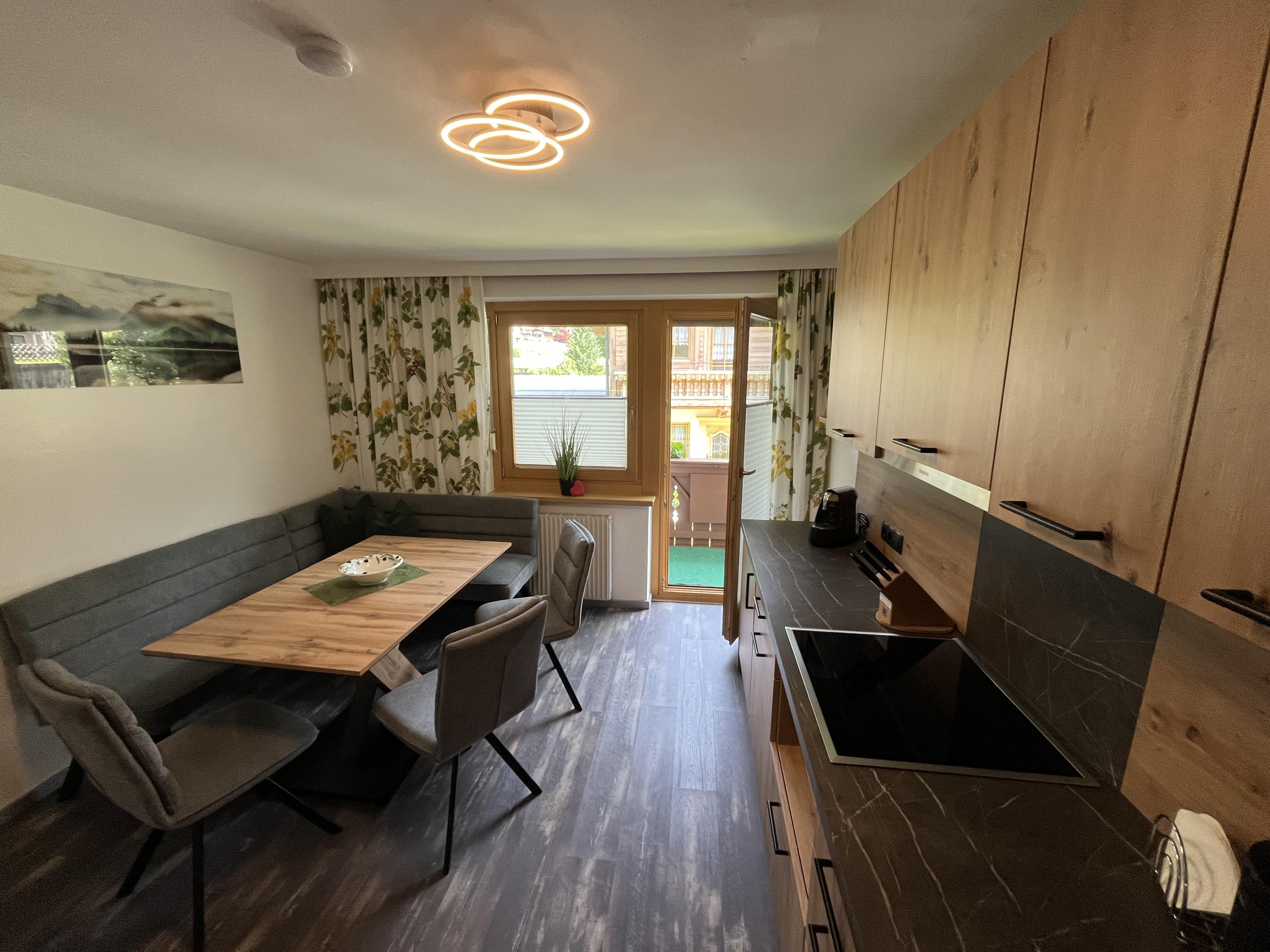 Wohnzimmer mit Esstisch, Stühlen, Couch, Fenster mit Blumenmuster-Vorhängen und moderner Deckenleuchte.