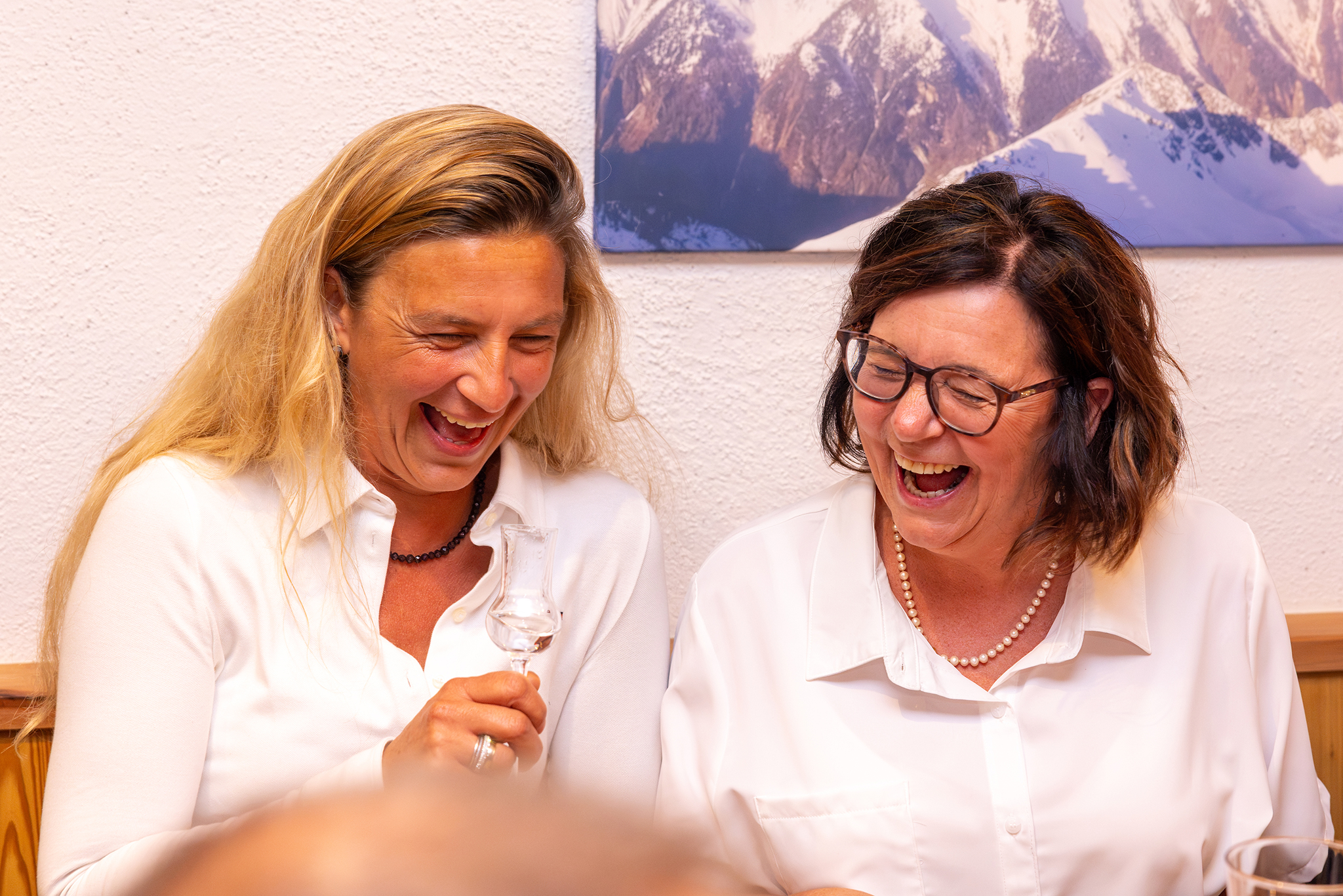 Zwei lachende Frauen in weißen Hemden sitzen Schulter an Schulter am Tisch, die blonde Frau links hält ein Schnapsglas vor der Brust, die Brünette mit Brille und Perlenkette neigt sich ihr lachend zu. An der Wand ein gerahmtes Bergpanorama.