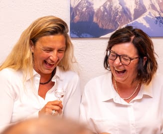 Zwei lachende Frauen in weißen Hemden sitzen Schulter an Schulter am Tisch, die blonde Frau links hält ein Schnapsglas vor der Brust, die Brünette mit Brille und Perlenkette neigt sich ihr lachend zu. An der Wand ein gerahmtes Bergpanorama.