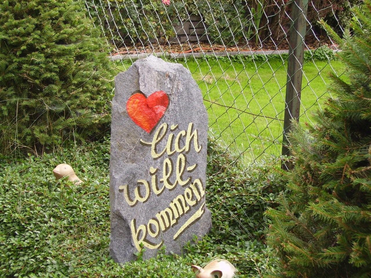 Stein mit Herz und Schriftzug 'lich will-bondmen' vor Gartenzaun, umgeben von Pflanzen.