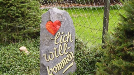 Stein mit Herz und Schriftzug 'lich will-bondmen' vor Gartenzaun, umgeben von Pflanzen.