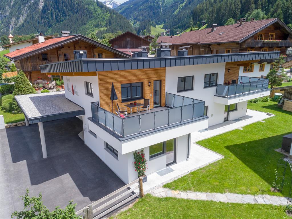 Modern multi-family house with balconies, surrounded by greenery and mountains, sunny day.
