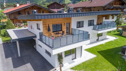 Modern multi-family house with balconies, surrounded by greenery and mountains, sunny day.