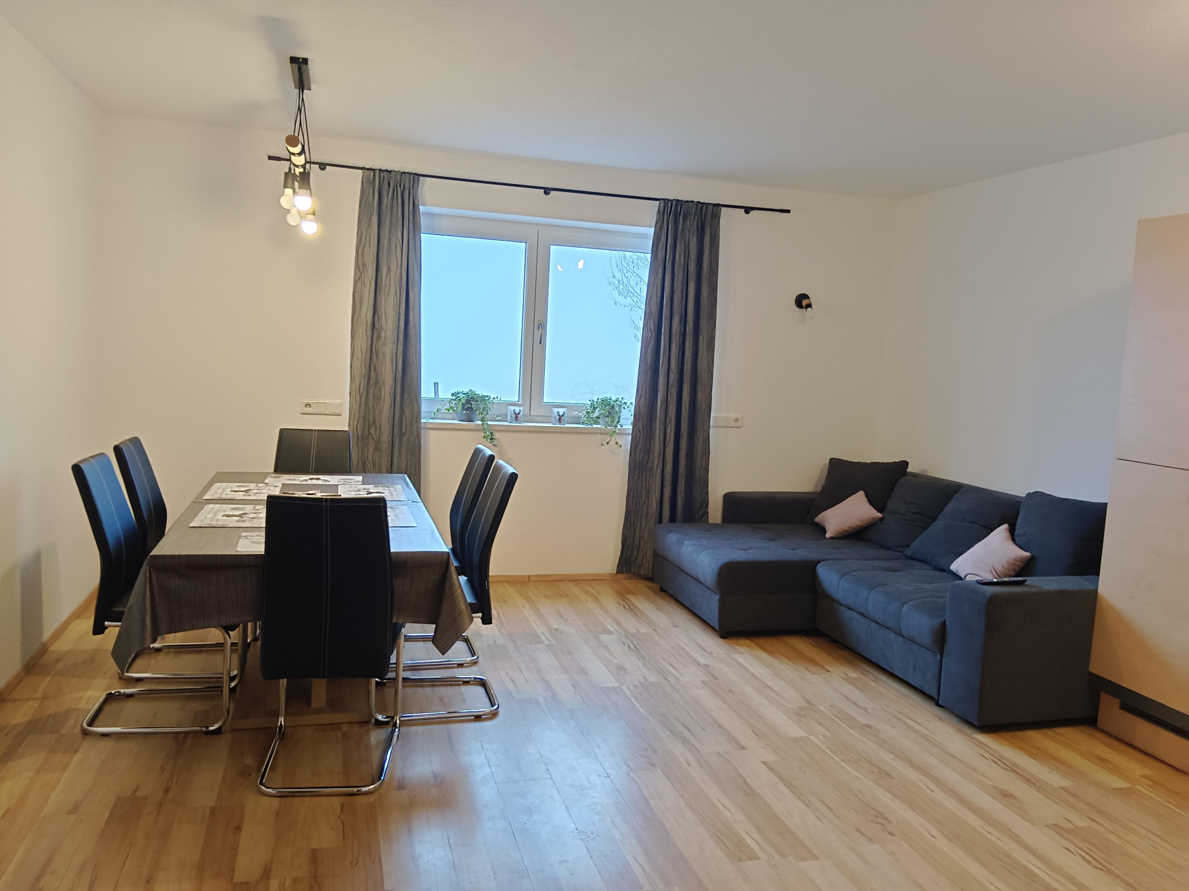Heller Esszimmer mit Esstisch, schwarzen Stühlen, Sofa und Fenster mit Vorhängen, Pflanzen und Deckenleuchte.