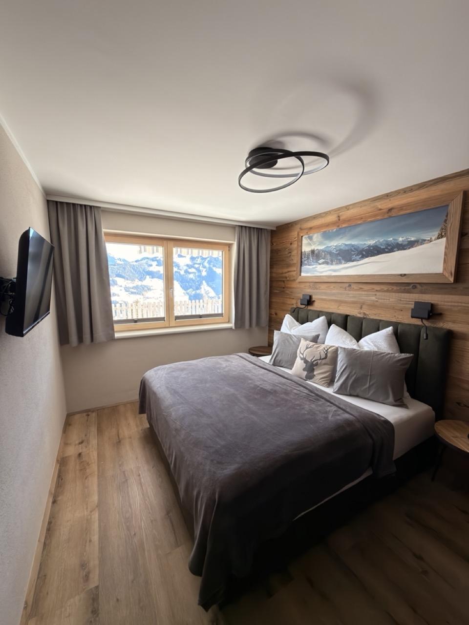 Schlafzimmer mit Bett, Holz-Wand, Fenster mit Bergblick, Wandlampe, TV an der Wand, und Deckenleuchte.