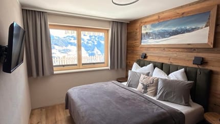 Schlafzimmer mit Bett, Holz-Wand, Fenster mit Bergblick, Wandlampe, TV an der Wand, und Deckenleuchte.