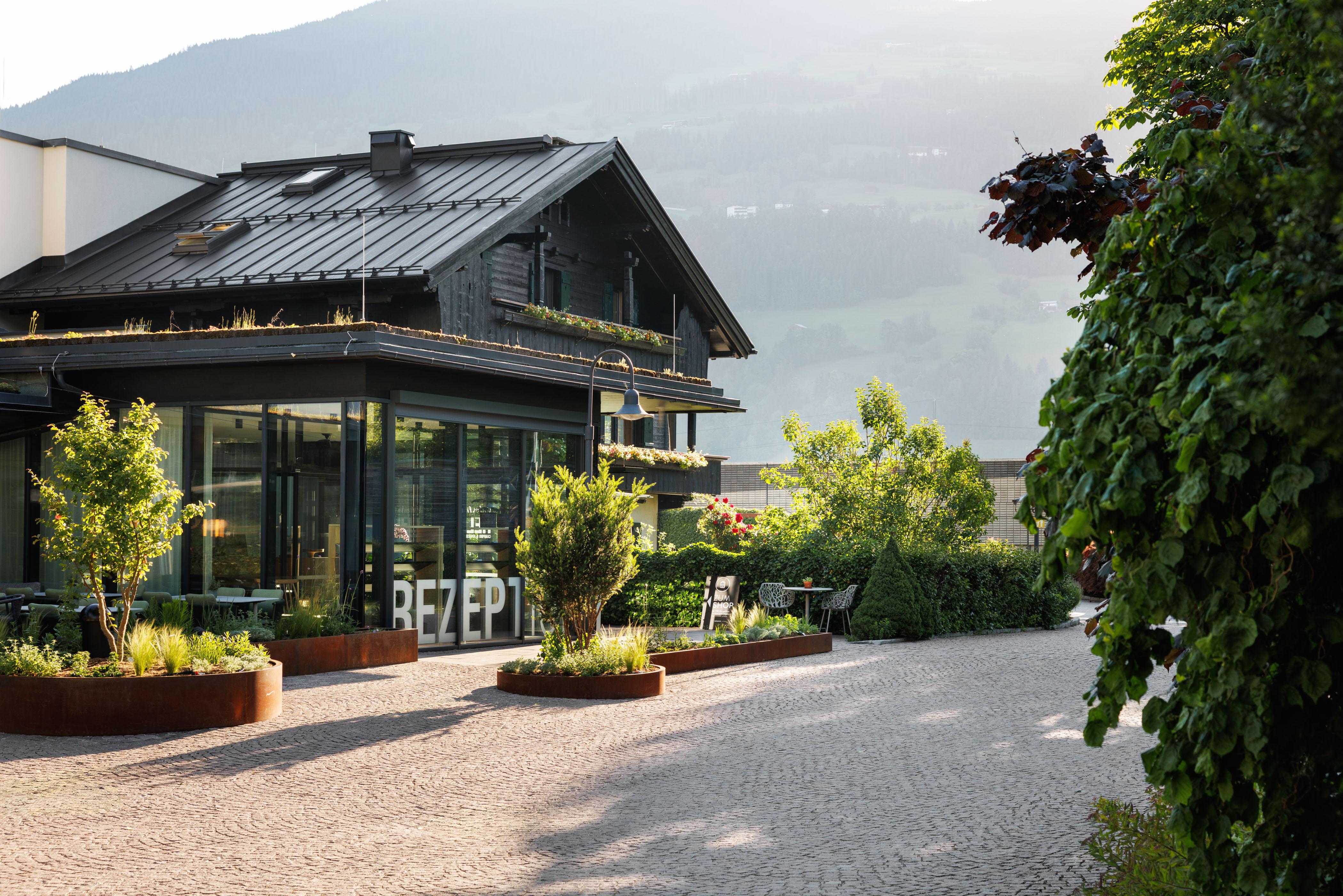 Modernes Haus mit dunklem Holz, große Fenster und umliegender Garten mit Pflanzen und Bäumen, im Hintergrund Berge.