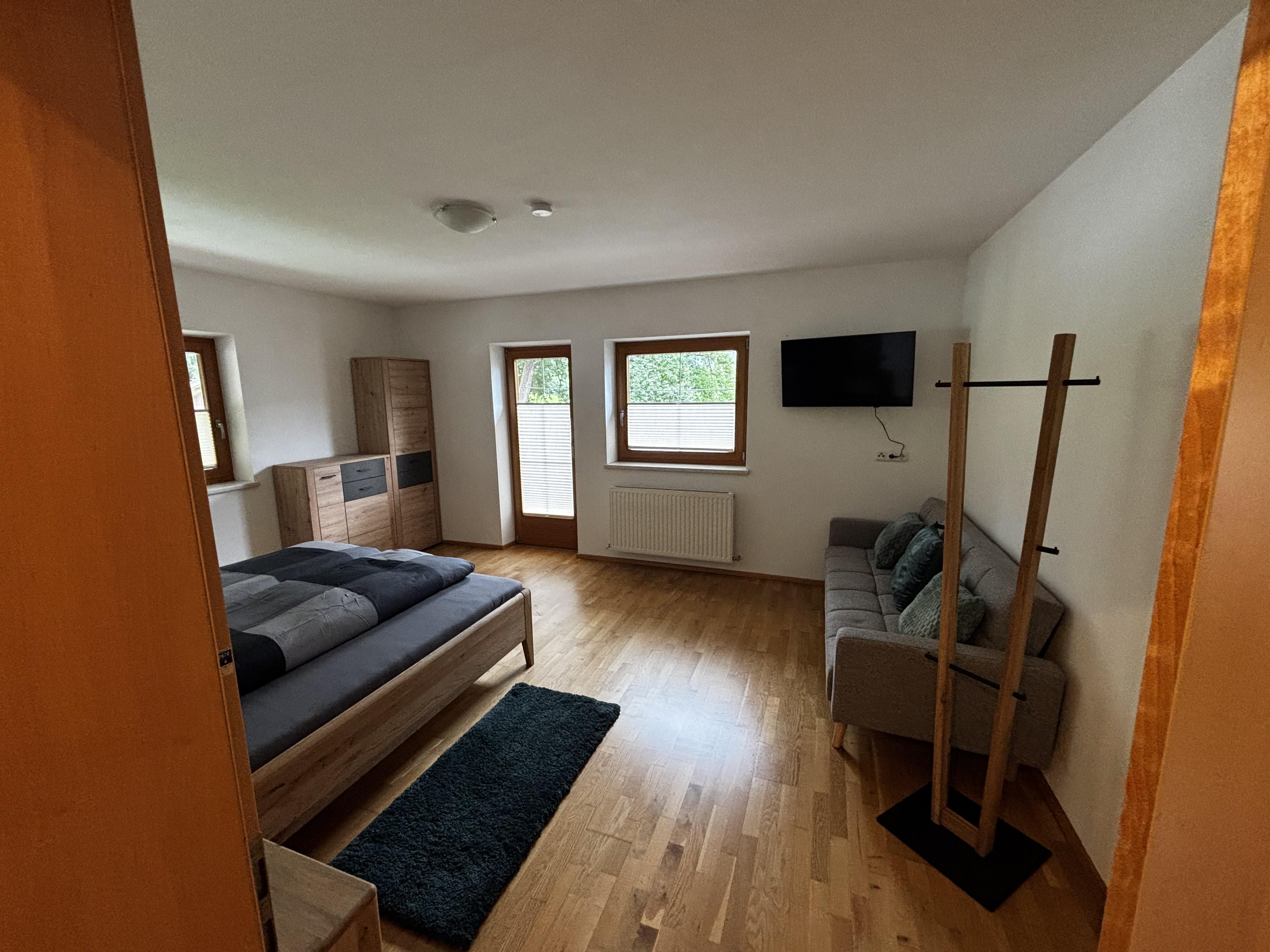 Innenaufnahme eines Schlafzimmers mit Bett, Sofa, TV und großen Fenstern, helle Holzböden, freundliche Atmosphäre.